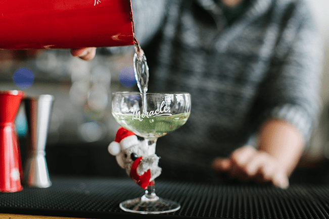 A small koala toy clinging to a martini glass, a bartender pours a drink in the background