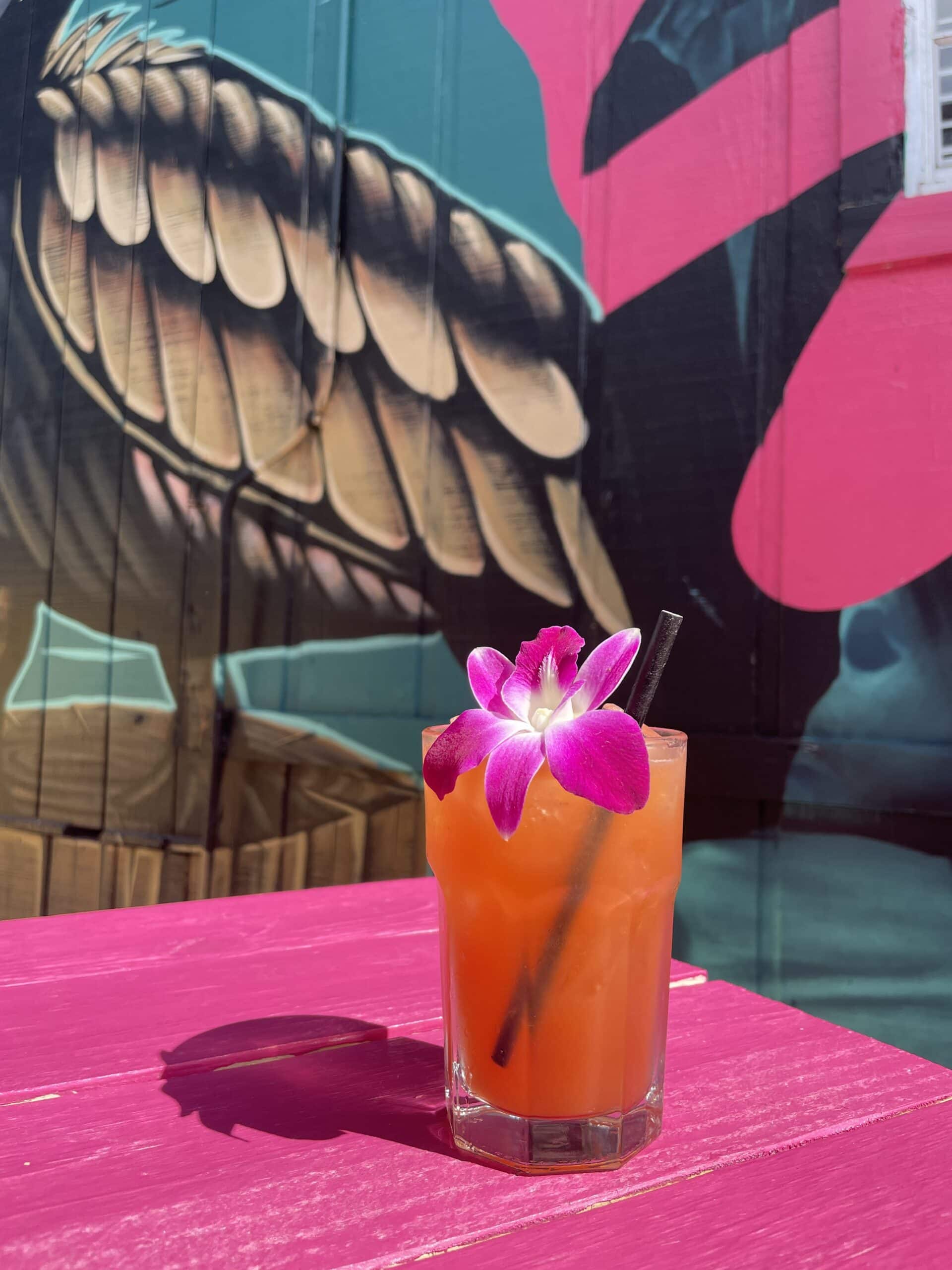 Image of red-colored cocktail in glass with purple orchid in front of colorful mural. 