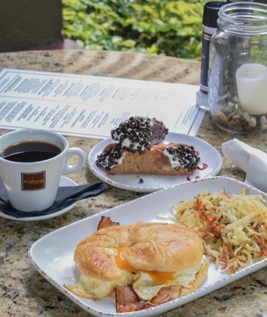coffee, a croissant sandwich, cannoli and hash browns arranged on a plate.