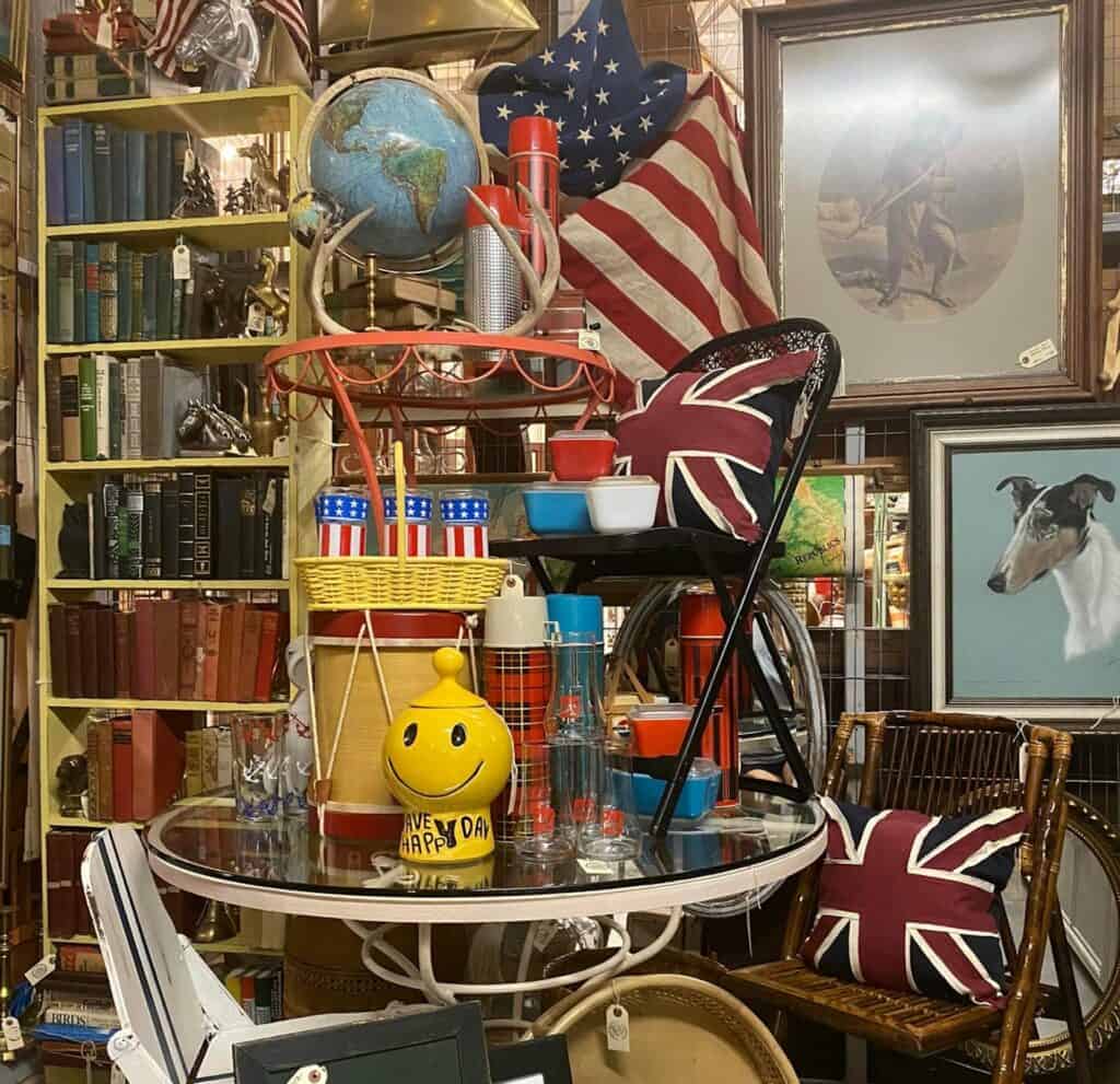 a circular glass table with a folding chair on top. A smiley face pepper shaker is at the center. A globe is set up next to an American flag and a United Kingdom Flag pillow. 